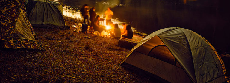 Mehrer Personen zelten am See und sitzen um ein Lagerfeuer herum