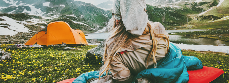 Eine blonde Frau liegt im Schlafsack auf einer wiese in den Bergen