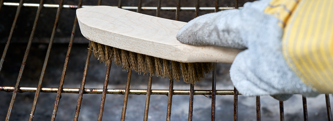 Die hölzerne Drahtbürste reinigt schmutzigen Grillrost. Lederschutzhandschuhe.