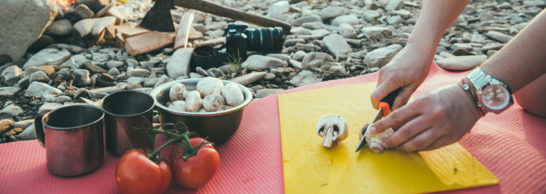 Camping kochen. Gemüse wird auf Brettchen geschnitten. Steine im Hintergrund.